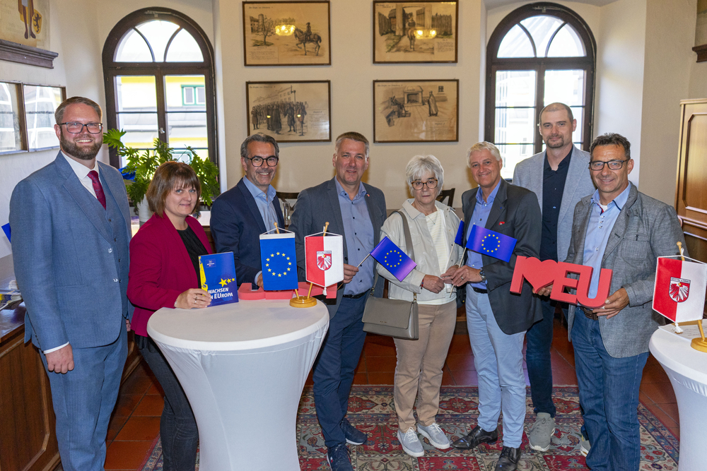 EU-Flaggen und ein Buch über Europa halten Personen in einem Raum mit historischen Bildern an der Wand.